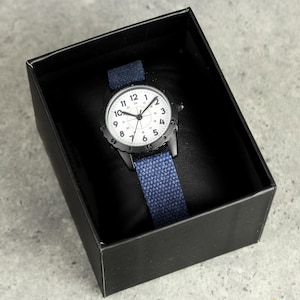 May include: A black wristwatch with a white face and blue canvas band is displayed inside a black box. The watch has black hands and hour markers. The band is a solid blue color.