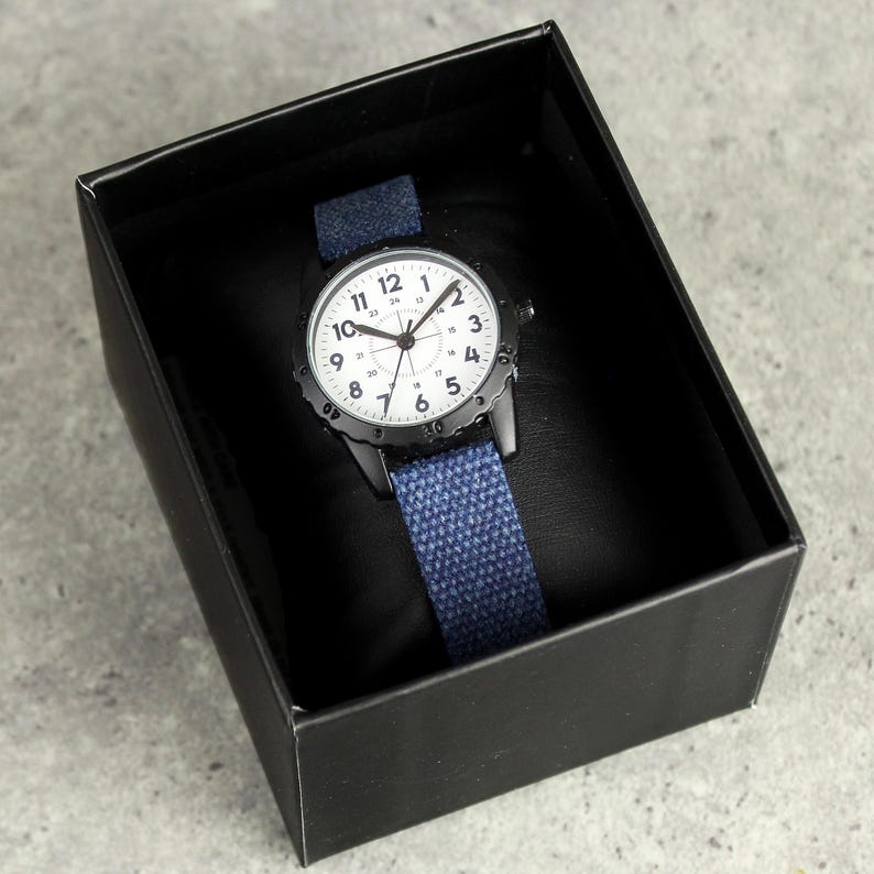 May include: A black wristwatch with a white face and blue canvas band is displayed inside a black box. The watch has black hands and hour markers. The band is a solid blue color.