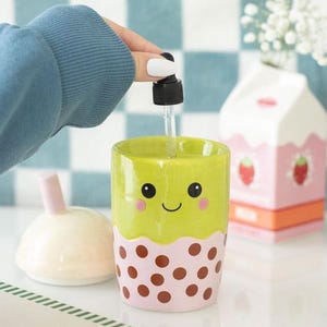 May include: A ceramic soap dispenser shaped like a bubble tea cup, featuring a light green top and a pink base with brown dots. The dispenser has a smiling face design and a black pump. A matching lid and a strawberry milk carton are also visible.