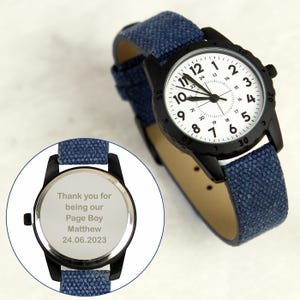 May include: A black wristwatch with a white face and blue canvas strap. The watch face has black numerals and hands. The back of the watch has engraved text: "Thank you for being our Page Boy Matthew 24.06.2023".