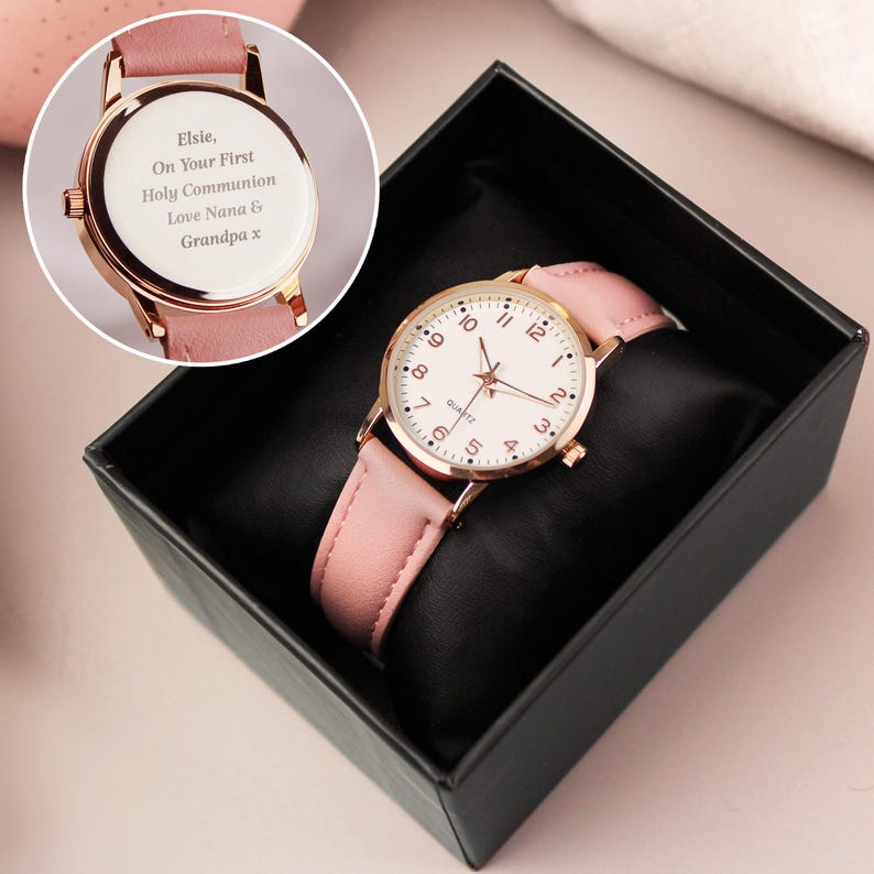 May include: A rose gold watch with a white face and pink leather band, presented in a black box. The watch face has black numerals and hands. The back of the watch is engraved with the text: "Elsie, On Your First Holy Communion Love Nana & Grandpa x."