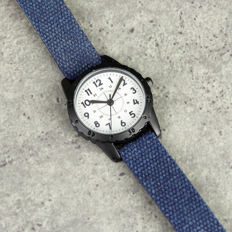 May include: A black wristwatch with a white face and blue canvas band. The watch face has black numerals and hands, with a second hand. The band is a solid blue color and is attached to the watch case.