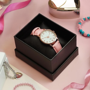 May include: A pink wristwatch with a white face and gold-tone accents rests in a black box. The watch has a pink leather band. Other jewelry and pink ribbons are in the background.