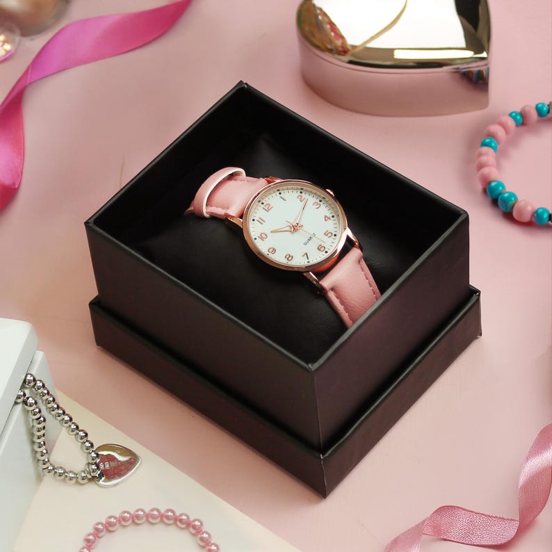 May include: A pink wristwatch with a white face and gold-tone accents rests in a black box. The watch has a pink leather band. Other jewelry and pink ribbons are in the background.