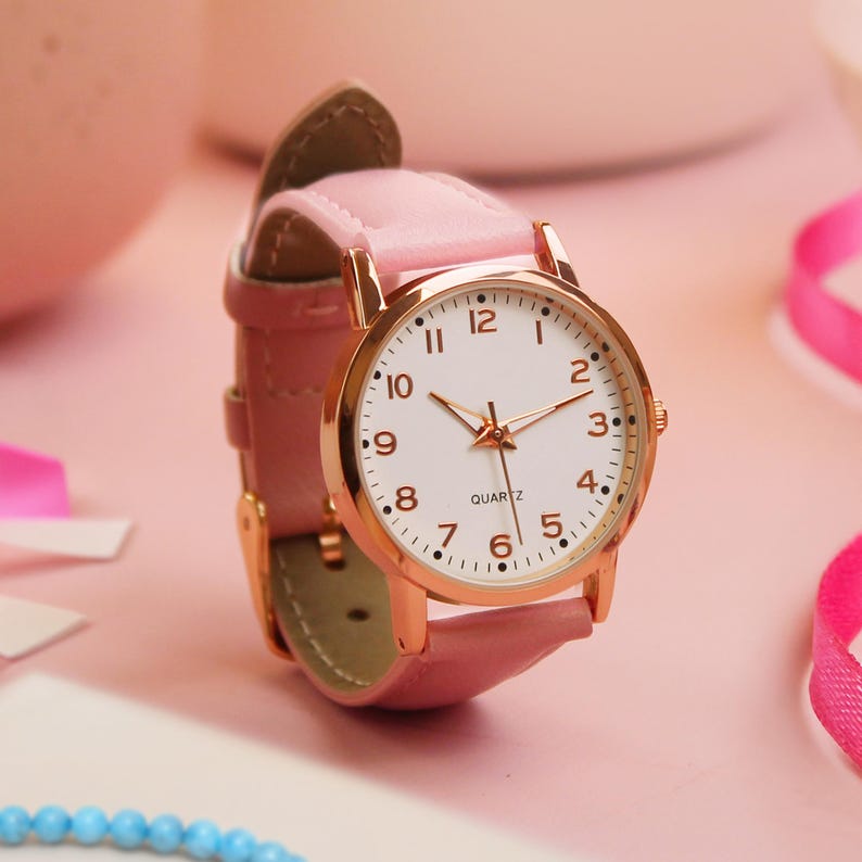 May include: A pink wristwatch with a gold-colored case and a white face. The watch has a pink leather band and a quartz movement. The watch face displays the numbers 1 through 12, with gold-colored hands.