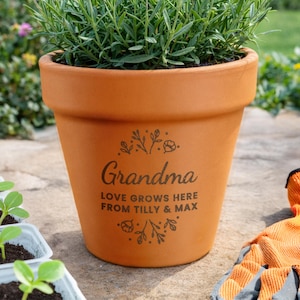 Könnte beinhalten: Ein Terrakotta-Blumentopf mit dem Wort "Oma" und dem Satz "LOVE GROWS HERE FROM TILLY & MAX" in Schwarz aufgedruckt. Der Topf ist mit grünen Pflanzen gefüllt und steht auf einer Steinoberfläche. Gartenhandschuhe sind im Hintergrund zu sehen.