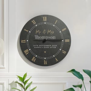 Op de afbeelding: Een ronde, zwarte wandklok met gouden Romeinse cijfers en wijzers. De klok heeft de tekst "Mr & Mrs Thompson" boven "15th September 2026 Happily Ever After". Een klein hartje staat eronder.