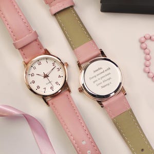 May include: Two pink leather strap watches with gold-tone cases. The watch faces are white with black numerals and hands. One watch has the text "Amelia, Every second with you is precious. Love Always, Nanny x" on the back.