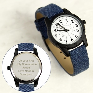 May include: A black wristwatch with a white face and blue denim strap. The watch face has black numerals and hands. The back of the watch has engraved text that reads "On your first Holy Communion Jacob Love Nana & Grandpa x."