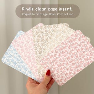 May include: Kindle clear case inserts in shades of blue, pink, and beige, adorned with a repeating bow and floral design. The inserts are held in a hand, part of the Coquette Vintage Bows Collection.