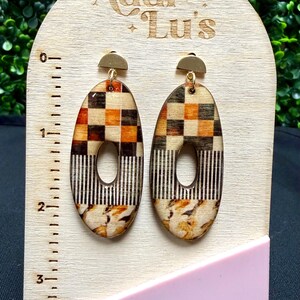 Peut inclure: Boucles d'oreilles ovales au design unique. Le haut présente un motif en damier marron, orange et crème. Le milieu a un motif à rayures verticales et le bas un motif floral. Les boucles d'oreilles sont exposées sur un présentoir en bois avec le texte "Addi + Lu's".