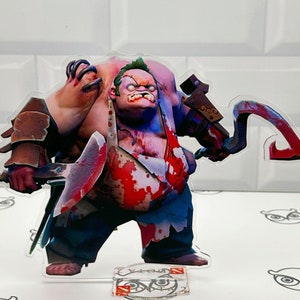 May include: A 3D printed figurine of a character from the video game Dota 2. The character is a large, muscular, green-skinned ogre with a red and white apron and a bloody hook. The figurine is standing on a clear acrylic base with the Dota 2 logo.