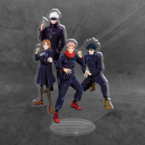 May include: A clear acrylic standee featuring four anime characters from Jujutsu Kaisen. The group includes a tall figure in a dark uniform with a blindfold, a person holding a hammer, a central figure with pink hair, and a crouching dark-haired figure.