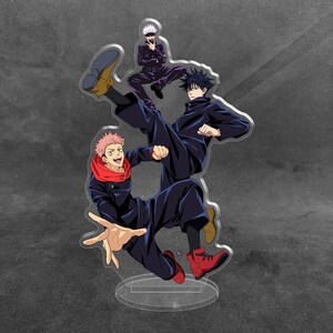 May include: A clear acrylic standee featuring three anime characters in dynamic poses. One character with pink hair wears a dark uniform and red scarf, another with dark hair kicks, and a third with white hair sits above them in a dark outfit.