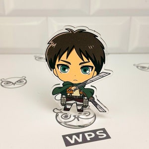 May include: A chibi-style acrylic stand featuring a character from the anime Attack on Titan. The character is holding a sword and has brown hair, green eyes, and a determined expression. The stand is clear and has a circular base.