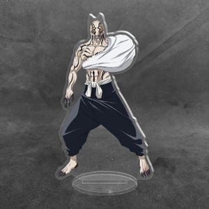 May include: A clear acrylic standee featuring a stylized anime character with a pale, mask-like face, dark markings on the torso and arms, and a white sling on the left arm. The figure wears dark trousers with a white sash and stands on a circular base.