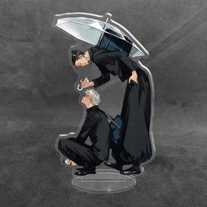 May include: A clear acrylic standee featuring two anime-style characters in black uniforms. One character with dark hair stands holding a white umbrella over a second character with white hair who is crouching on the ground. Both carry dark backpacks.