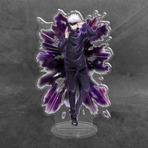 May include: An acrylic standee featuring a character with white hair and a black blindfold, wearing a dark blue high-collared jacket and matching trousers. The character is posed against a vibrant purple and black burst effect background.