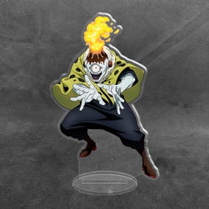 May include: A clear acrylic standee featuring a one-eyed character with a volcanic head erupting with yellow and orange flames. The figure wears a yellow cape with black spots over dark pants and brown boots, posed with hands outstretched forward.