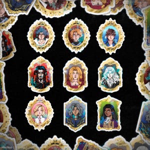 May include: A selection of stickers displaying portraits of diverse characters within decorative gold frames. Each portrait presents a different individual with distinct hairstyles and clothing, set against vibrant backgrounds. The stickers have a glossy finish and are arranged on a black surface.
