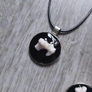 Puede incluir: Un collar con colgante redondo con fondo negro y dise&ntilde;o blanco en forma de hueso. El colgante est&aacute; sujeto a un collar de cord&oacute;n negro con cierre plateado. El collar se muestra sobre una superficie de madera gris.
