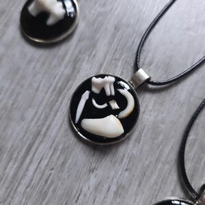 Puede incluir: Collar con colgante redondo con fondo negro y borde plateado. El colgante presenta un dise&ntilde;o blanco con tem&aacute;tica dental. El collar tiene un cord&oacute;n negro. Hay colgantes adicionales visibles en el fondo.