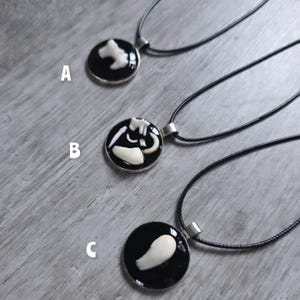 Puede incluir: Tres collares con colgantes negros y plateados con amuletos blancos en forma de hueso. Cada colgante es redondo con un fondo negro y un bisel plateado. Los collares se exhiben sobre una superficie de madera gris claro.