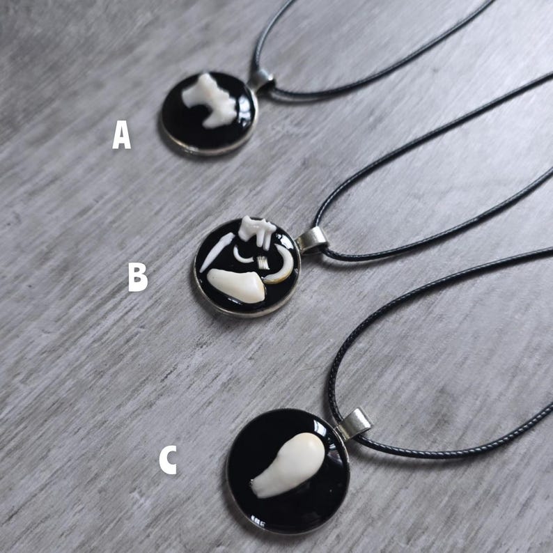 Puede incluir: Tres collares con colgantes negros y plateados con amuletos blancos en forma de hueso. Cada colgante es redondo con un fondo negro y un bisel plateado. Los collares se exhiben sobre una superficie de madera gris claro.
