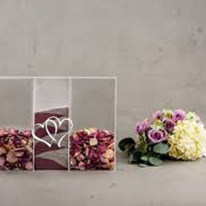 Könnte beinhalten: Drei transparente Acrylboxen mit einem Herzdesign auf der Vorderseite. Jede Box ist mit rosa und weißen Blütenblättern gefüllt. Im Vordergrund befindet sich ein Strauß aus lila und weißen Blumen.