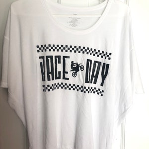 May include: White t-shirt with a black graphic that says "Race Day" with a silhouette of a person riding a dirt bike. The graphic is surrounded by a black and white checkered pattern.