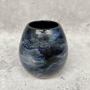 May include: A small, round ceramic vase with a dark blue and black marbled glaze. The vase has a wide opening and a smooth, glossy finish. The colour transitions from dark blue to black, creating a unique, artistic look. This decorative vase is suitable for home decor.