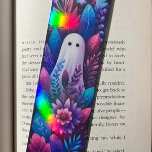 Op de afbeelding: Rechthoekige boekenlegger met een witte geest omringd door kleurrijke bladeren en bloemen. De boekenlegger heeft een holografisch regenboogeffect. Het nummer 85 is zichtbaar aan de onderkant.