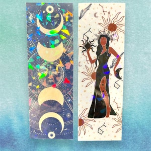 May include: Two holographic bookmarks with celestial designs. One bookmark features a dark blue background with gold crescent moons and stars. The other bookmark features a white background with a gold silhouette of a woman holding a sun and surrounded by stars and suns.