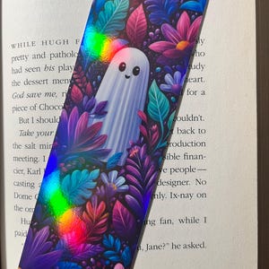 Op de afbeelding: Een rechthoekige boekenlegger met een schattige spookillustratie omringd door kleurrijke bloemen en bladeren. Het ontwerp bevat tinten paars, roze, blauw en groen met een holografisch effect. De boekenlegger ligt op een open boek.