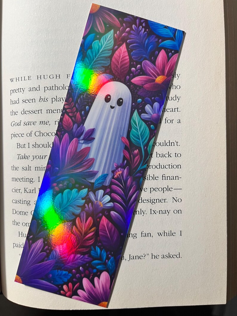 Op de afbeelding: Een rechthoekige boekenlegger met een schattige spookillustratie omringd door kleurrijke bloemen en bladeren. Het ontwerp bevat tinten paars, roze, blauw en groen met een holografisch effect. De boekenlegger ligt op een open boek.