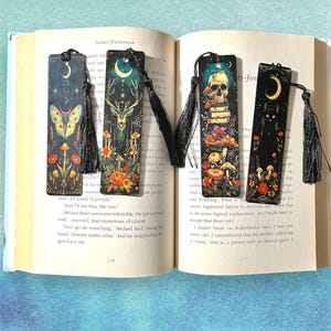 May include: Set of five bookmarks with black tassels. Each bookmark features a different design with a dark background and colorful accents. The designs include a moth, a deer, a skull, a cat, and mushrooms.