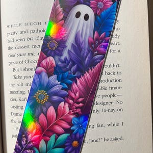 Op de afbeelding: Rechthoekige boekenlegger met een wit spookontwerp, tegen een achtergrond van paarse, roze en blauwe bloemen en bladeren. De boekenlegger heeft een holografisch effect en is bedoeld voor boeken.