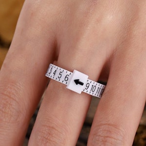 May include: White plastic ring sizer with black numbers and an arrow pointing to the right. The sizer is on a finger, showing sizes 3 through 11.
