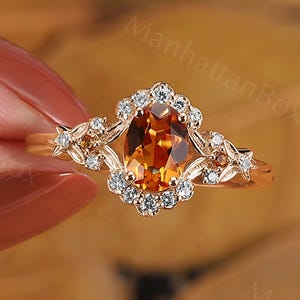 May include: A gold ring with a pear-cut, orange gemstone, encircled by small, clear stones. The ring has a floral design with leaves and small clear stones on the band. The ring is set against a blurred backdrop of autumn leaves.