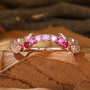Puede incluir: Un anillo de oro rosa con un diseño floral. El anillo presenta pequeñas piedras preciosas rosas y magenta engastadas en una banda curva. El anillo está sobre un lecho de hojas secas.