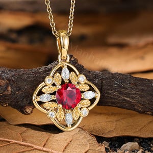 May include: A gold pendant necklace featuring a central round-cut red gemstone surrounded by a decorative frame of gold leaves, small marquise-cut white stones, and round white stones, all suspended from a delicate gold chain link necklace.
