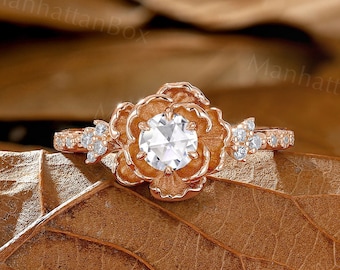 May include: A rose gold engagement ring with a round center stone and smaller stones set in a floral design. The ring is on a bed of brown leaves.