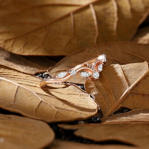 K&ouml;nnte beinhalten: Ein Ring aus Ros&eacute;gold mit einem zarten, geschwungenen Design und kleinen, klaren Edelsteinen. Der Ring liegt auf einem Hintergrund aus braunen Bl&auml;ttern.