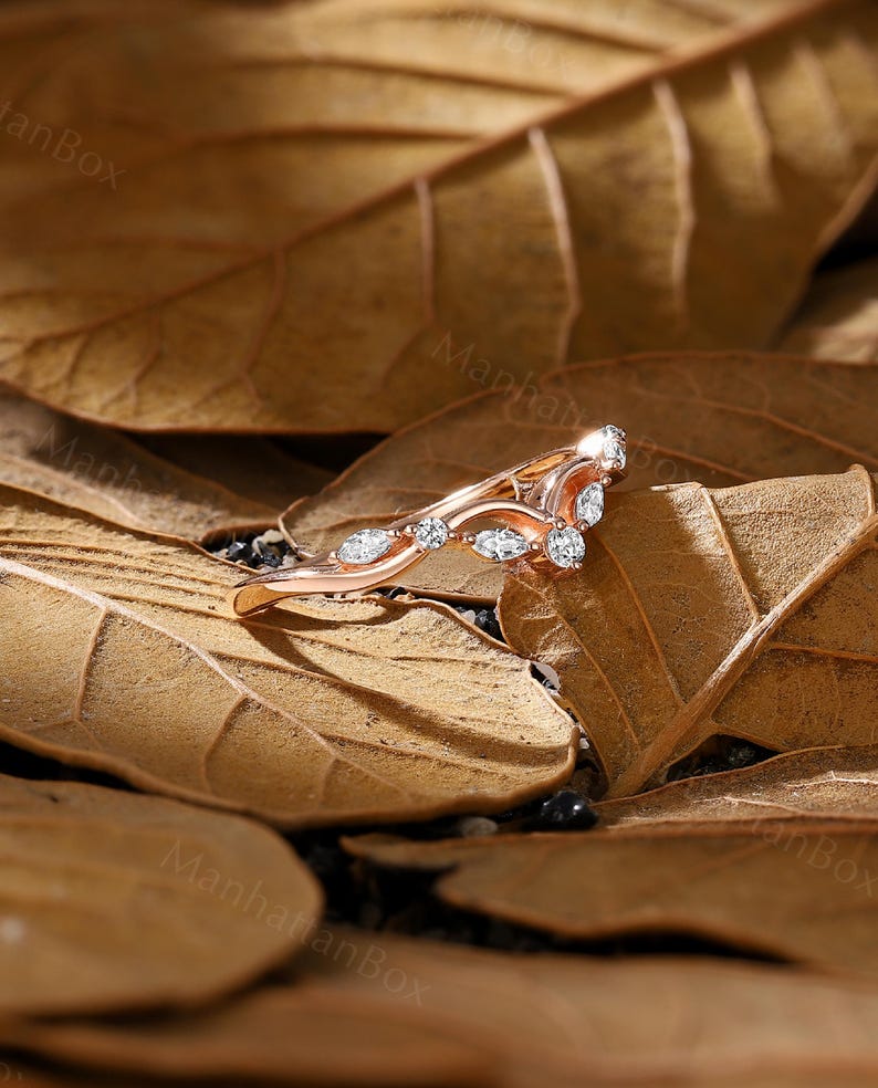 K&ouml;nnte beinhalten: Ein Ring aus Ros&eacute;gold mit einem zarten, geschwungenen Design und kleinen, klaren Edelsteinen. Der Ring liegt auf einem Hintergrund aus braunen Bl&auml;ttern.