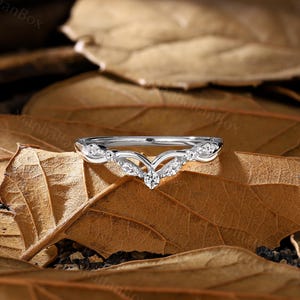 K&ouml;nnte beinhalten: Ein zarter Silberring mit einem V-f&ouml;rmigen Design und kleinen funkelnden Steinen. Der Ring liegt auf einem Hintergrund aus braunen Bl&auml;ttern.