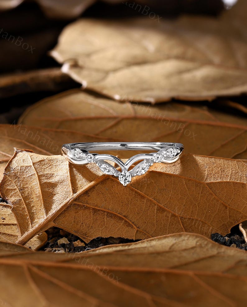 K&ouml;nnte beinhalten: Ein zarter Silberring mit einem V-f&ouml;rmigen Design und kleinen funkelnden Steinen. Der Ring liegt auf einem Hintergrund aus braunen Bl&auml;ttern.