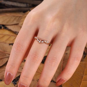 K&ouml;nnte beinhalten: Ein zarter Ring aus Ros&eacute;gold mit einem geschwungenen Design und kleinen, klaren Steinen. Der Ring befindet sich an einer Hand mit braunem Nagellack.