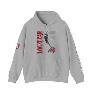 Puede incluir: Una sudadera con capucha gris con un gráfico rojo y blanco de una gimnasta en el aire. El gráfico incluye el texto "LOCKLEAR" y "MIDN". Las mangas tienen el texto "LOCKLEAR" impreso verticalmente en rojo.