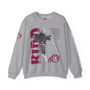 May include: A heather gray sweatshirt featuring a skier in mid-air, wearing a cowboy hat. The design includes the text "KID" in maroon, a quote, and the words "All-Stars". The sleeve has the text "nullalistars.com".