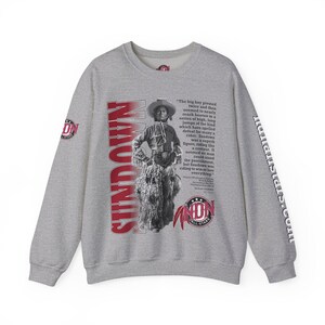 Puede incluir: Sudadera gris con un estampado gráfico rojo y blanco de un vaquero montando un caballo salvaje. El texto "Sundown" está impreso verticalmente en el lado izquierdo de la imagen. El texto "All-Stars" está impreso en rojo en la esquina inferior derecha de la imagen. El texto "The big bay pivoted twice and then seemed to nearly reach heaven in a series of high, long jumps of the kind which have spelled defeat for many a rider. Sundown was a superb figure, riding like a centaur. It seemed no man could stand the punishment, but Sundown was riding to win or lose everything." está impreso en negro en el lado derecho de la imagen.